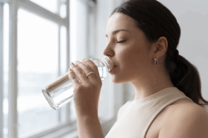 Closeup of woman drinking water from a glass bottle in front of a window.