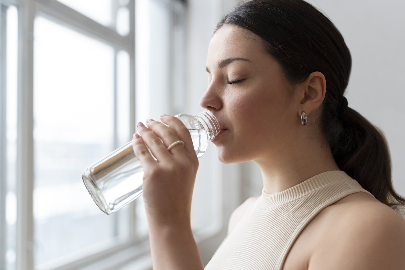 Closeup of woman drinking water from a glass bottle in front of a window.