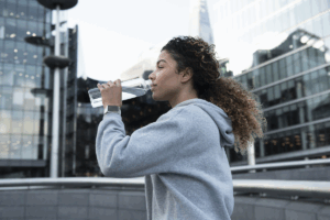 Young woman in a hoodie drinking water while walking in a city center.