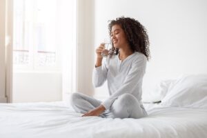 Woman sitting on a bed, drinking a glass of water as she starts her day.