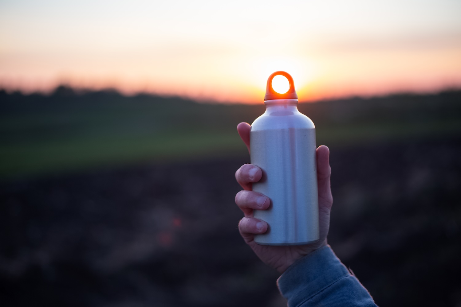 Person holding an aluminum water bottle in front of the sun as it's setting.