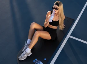 Blonde woman drinking One Water while sitting on a tennis court.