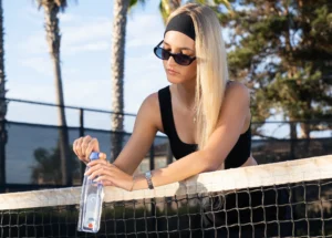 Woman opening a One Water bottle while leaning on a tennis net.